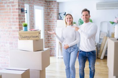 Genç ve güzel bir çift, yeni evlerinde karton kutuların etrafında durup kameraya hayrete düşmüş ve gülümserken elleri ve parmaklarıyla işaret ediyorlar..