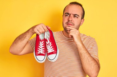 Elinde kırmızı spor ayakkabıları olan genç bir adam izole edilmiş sarı arka planda dikiliyor ciddi bir yüz soru düşünüyor, çok karışık bir fikir.