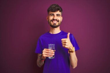 Dövmeli genç adam, izole edilmiş mor arka planda duran su bardağını içerken mutlu bir şekilde gülümsüyor.