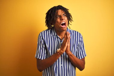 Rastgele çizgili tişört giyen Afro bir adam izole edilmiş sarı arka plan üzerinde yalvarıyor ve dua ediyor. Yüzünde umut ifadesi olan çok duygusal ve endişeli bir ifade var. Af diliyor. Din kavramı.