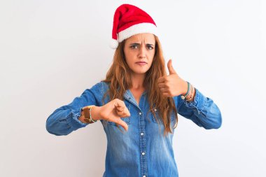 Genç ve güzel kızıl saçlı bir kadın, izole edilmiş arka planda noel şapkası takıyor. Baş parmak işareti yapıyor, anlaşmazlık ve uzlaşma ifadesi takınıyor. Çılgın çatışma