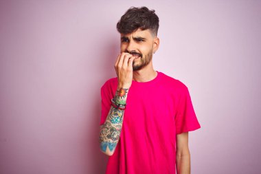 İzole edilmiş pembe arka plan üzerinde duran dövmeli genç adam elleri ağız yiyen tırnaklarla gergin ve stresli görünüyor. Endişe sorunu.