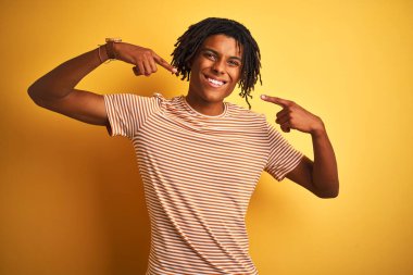 Rastalı tişörtlü Afro adam izole edilmiş sarı arka planda dikiliyor, gülümsüyor ve parmakları, dişleri ve ağzıyla işaret ediyor. Diş sağlığı konsepti.