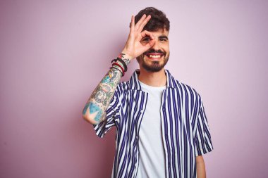 Dövmeli, çizgili tişörtlü genç adam pembe arka planda dikiliyor. El hareketleriyle gülümsüyor ve mutlu yüzüyle parmaklarının arasından bakıyor..