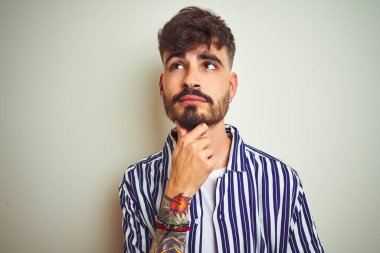 İzole edilmiş beyaz arka plan üzerinde duran çizgili tişörtlü genç bir adam çenesinde elle soru soran, düşünceli bir ifade. Düşünceli bir yüzle gülümsüyor. Şüphe kavramı.