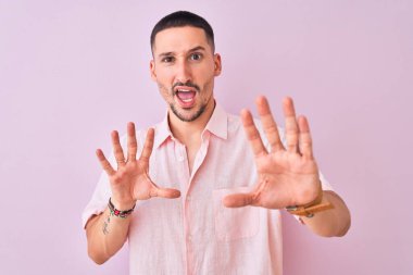 Pembe gömlekli genç adam izole edilmiş arka planda duruyor korku ifadesinden korkuyor ve dehşete düşüyor. El hareketlerini durdur, şok içinde bağır. Panik konsepti.