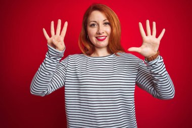 Kızıl saçlı genç bir kadın donanma gömleği giyiyor. Kırmızı izole edilmiş arka plan üzerinde duruyor ve dokuz numarayı işaret ediyor. Kendinden emin ve mutlu bir şekilde gülümsüyor..