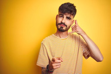 İzole edilmiş sarı arka plan üzerinde duran çizgili tişörtlü genç adam gülümsüyor telefonda konuşuyor ve seni işaret ediyor. Ara beni..