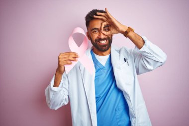 Kanser kurdelesi tutan genç Hintli doktor mutlu yüzüyle pembe arka planda dikiliyor.