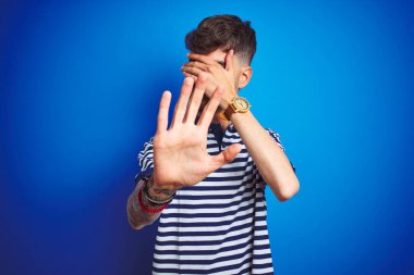 Çizgili polo giyen genç adam izole edilmiş mavi arka planda duruyor gözleri ellerle örtüyor ve üzgün ve korku ifadeleriyle dur hareketi yapıyor. Utanç verici ve olumsuz bir kavram..