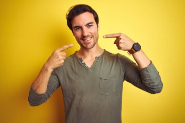 İzole edilmiş sarı arka planda dikilen günlük tişört giyen genç yakışıklı adam neşeli bir şekilde gülümseyip parmaklarını ve ağzını işaret ediyor. Diş sağlığı konsepti.