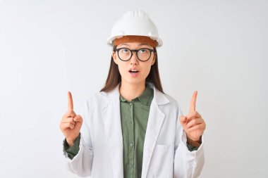 Genç Çinli mühendis kadın kaban gözlüğü takıyor beyaz arka planda şaşkın şaşkın yukarı bakıyor ve parmaklarıyla işaret ediyor ve kollarını kaldırıyor..