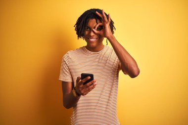 Rastalı saçlı Afro-Amerikalı adam, izole edilmiş sarı arka plan üzerinde akıllı telefon kullanıyor. Gülümsüyor, gülümsüyor.