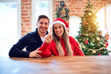 Noel Baba şapkası takan genç bir çift evde Noel ağacının etrafında oturmuş gülümseyen, kendinden emin ve mutlu bir şekilde bir numarayı işaret ediyor..