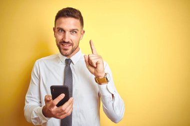 Genç ve yakışıklı bir iş adamı sarı arka plan üzerinde akıllı telefon kullanıyor. Bir fikir veya soru işareti ile şaşırmış, mutlu bir yüz, bir numara.