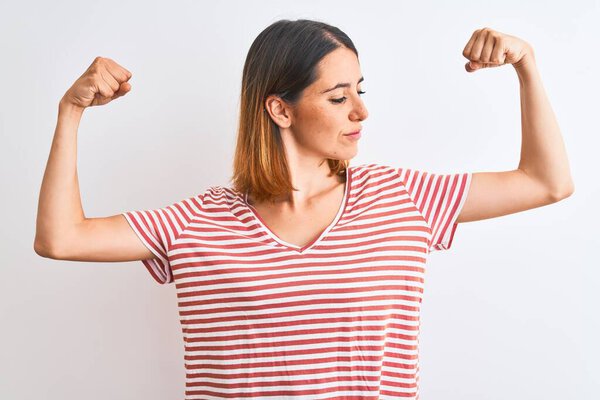 Красивая рыжеволосая женщина в обычной полосатой красной футболке на изолированном заднем плане, показывающая мышцы рук, гордо улыбающиеся. Фитнес-концепция
.