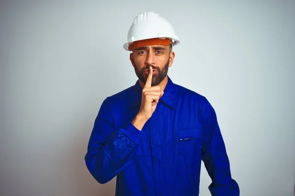 Handsome indian worker man wearing uniform and helmet over isolated white background asking to be quiet with finger on lips. Silence and secret concept.