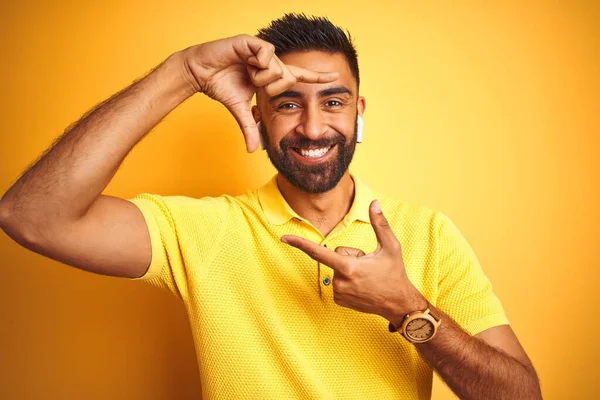Young indian man listening to music using earphones standing over isolated yellow background smiling making frame with hands and fingers with happy face. Creativity and photography concept.