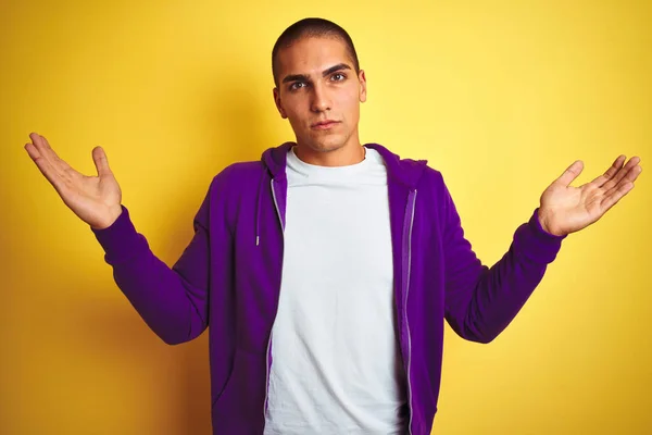 黄色の隔離された背景に紫色のスウェットシャツを着た若いハンサムな男は 腕と手で提起した混乱した表情 疑いの概念 — ストック写真