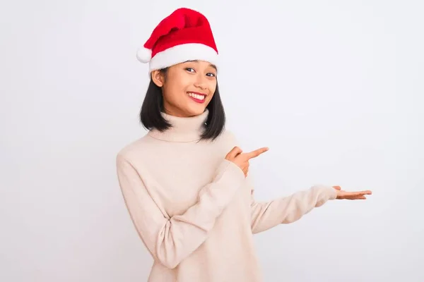 若いです美しい中国の女性身に着けているクリスマスのサンタの帽子隔離された白い背景に驚いて 手で提示しながら 指で指してカメラに笑顔 — ストック写真