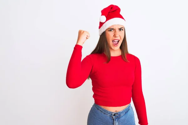 孤立した白い背景の上に立っているクリスマスサンタの帽子を身に着けている若い美しい女の子は怒っていると怒って叫んでいる間拳をフラストレーションと激怒を上げる 激怒と積極的な概念 — ストック写真