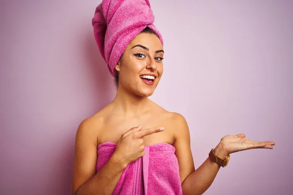 孤立したピンクの背景にシャワーの後にタオルを身に着けている若い美しい女性は驚きと手で提示しながら カメラに笑顔と指で指して — ストック写真