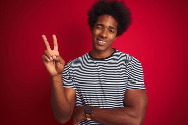 Afro saçlı genç Amerikalı adam izole edilmiş kırmızı arka plan üzerinde deniz mavisi çizgili tişört giyiyor ve kameraya gülümsüyor zafer işareti yapıyor. İki numara..