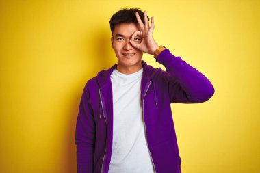 Mor kazak giyen Asyalı genç adam izole edilmiş sarı arka planda duruyor. El hareketleriyle gülümsüyor ve mutlu yüzle parmaklarının arasından bakıyor..
