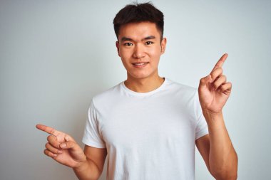 İzole edilmiş beyaz arka planda duran tişört giyen Asyalı genç adam parmaklarıyla farklı yönleri gösteren kendine güvenle gülümsüyor. Reklam için boşluk kopyala