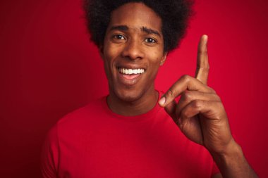 Afro saçlı genç Amerikalı adam izole edilmiş kırmızı arka planda dikilen tişörtlü bir fikir ya da soru işareti ile şaşırdı.