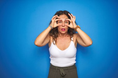 Kıvırcık saçlı orta yaşlı bir kadın mavi izole edilmiş arka planda ayakta duruyor parmaklarıyla gözlerini açmaya çalışıyor, uykulu ve yorgun bir şekilde sabah yorgunluğu çekiyor.