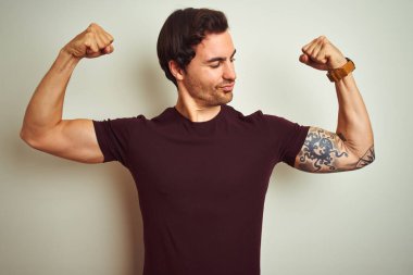 İzole edilmiş beyaz arka planda mor bir tişört giyen yakışıklı genç adam gururla gülümseyen kol kaslarını gösteriyor. Fitness konsepti.