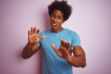 Afro saçlı, mavi tişörtlü, izole edilmiş pembe arka planda duran, korku ifadesinden korkan ve dehşete düşmüş Afro-Amerikan bir adam. El hareketini durdur, şok içinde bağır. Panik konsepti.