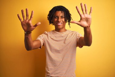 Rastalı tişörtlü Afro adam izole edilmiş sarı arka plan üzerinde duruyor ve parmaklarını on numarayı gösterirken kendinden emin ve mutlu gülümsüyor..
