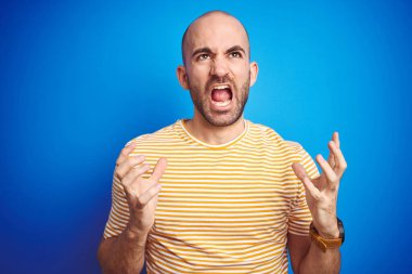 Mavi yalıtılmış arka plan üzerine gündelik tişört giyen sakallı genç kel bir adam sinirli bir şekilde bağırıyor ve agresif bir ifade ve kolla bağırıyor. Hayal kırıklığı konsepti.