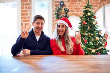 Noel Baba şapkası takan genç bir çift evdeki Noel ağacının etrafında oturmuş zafer işareti yaparken gülümsüyor. İki numara..