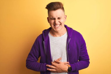 Mor eşofman üstü giyen genç yakışıklı adam midesi üzerinde, izole edilmiş sarı arka plan üzerinde duruyor çünkü hazımsızlık, acı verici bir hastalık kendini iyi hissetmiyor. Ache kavramı.