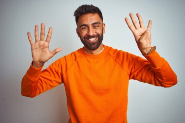 İzole edilmiş beyaz arka planda turuncu süveter giyen genç Hintli adam dokuz numaralı parmaklarıyla işaret ederken kendinden emin ve mutlu gülümsüyor..