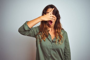 Yeşil tişörtlü genç bir kadın, gri yalıtılmış arka planda dikiliyor. Şok olmuş bir şekilde yüzünü ve gözlerini kapıyor. Utanmış bir ifadeyle parmaklarının arasından bakıyor..