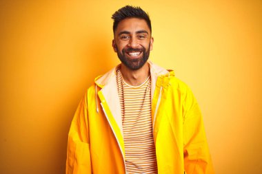 Yağmurluk giyen genç Hintli adam izole edilmiş sarı arka plan üzerinde mutlu ve serin bir gülümseme ile duruyor. Şanslı kişi..