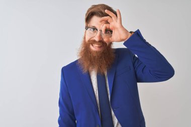 Genç kızıl saçlı, takım elbiseli ve gözlüklü bir iş adamı izole edilmiş beyaz arka planda el hareketleriyle gülümsüyor ve mutlu yüzle parmaklarının arasından bakıyor..