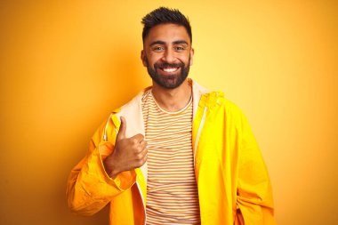 Sarı arka planda duran yağmurluk giyen genç Hintli adam mutlu bir el hareketi yapıyor. Kameraya bakıp başarıyı gösteren ifade onaylanıyor.