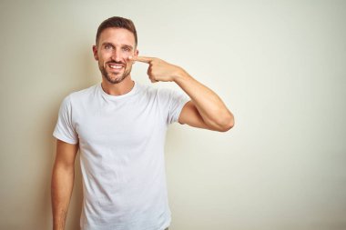 İzole edilmiş arka plan üzerine günlük beyaz tişört giyen genç yakışıklı bir adam. El parmağı ve burunla işaret ediyor. Neşeli bir şekilde gülümsüyor. Güzellik konsepti