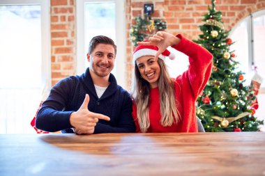 Noel Baba şapkası takan genç bir çift evde Noel ağacının etrafında oturmuş gülümsüyor ve mutlu yüzlü eller ve parmaklarla çerçeve yapıyor. Yaratıcılık ve fotoğrafçılık kavramı.