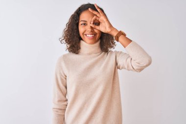 Balıkçı yaka kazak giyen genç bir kadın izole edilmiş beyaz arka planda dikiliyor. El hareketleriyle gülümsüyor, gözleriyle mutlu yüzle parmaklarının arasından bakıyor..