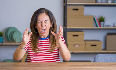 Orta yaş yaşlı kadın evde masada oturan kızgın ve öfkeli öfke ile bağırırken sinirli ve öfkeli yumruklar yükselterek. Öfke ve agresif kavram.