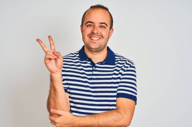 Sıradan çizgili polo giyen genç bir adam izole edilmiş beyaz arka planda durup kameraya gülümseyen ve parmaklarıyla zafer işareti yapan mutlu bir yüzle. İki numara..