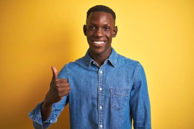 Kotlu tişört giyen genç Afro-Amerikan bir adam izole edilmiş sarı arka planda duruyor ve elleriyle mutlu bir jest yapıyor. Kameraya bakıp başarıyı gösteren ifade onaylanıyor.