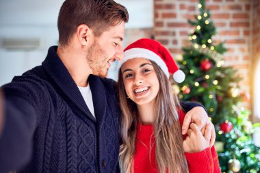 Mutlu ve kendinden emin gülümseyen genç bir çift. Evdeki Noel ağacının etrafında kamerayla selfie çekmek.
