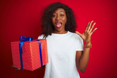 Genç Afrikalı Amerikalı bir kadın elinde doğum günü hediyesi ile kırmızı arka planda mutlu ve heyecanlı duruyor. Kazanan ifade zaferi büyük bir gülümsemeyle kutluyor ve ellerini kaldırıyor.